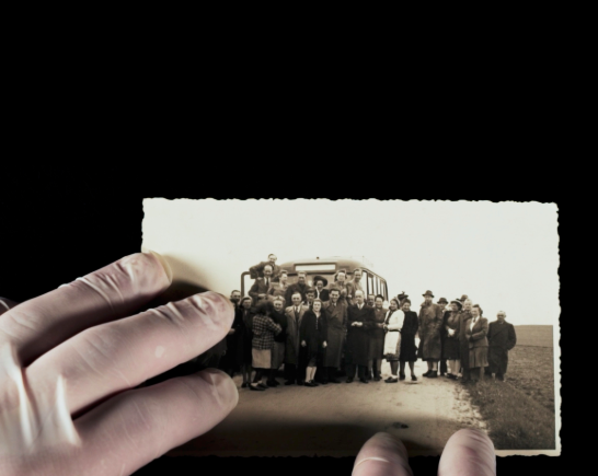 Two hands placed on the edge of a black and white photo of a group of people smiling and posing for a group photo in front of a bus used to transport disabled people to Nazi Death Camps
