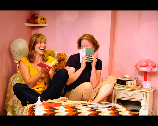 Two women sit together on a bed with a plaid blanket, reading books and laughing