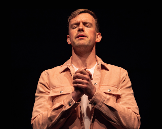 Irish actor Tom Moran on stage with hands clasped together in a ‘Big Fat Filthy Disgusting Liar’ in North American tour - Photo by Owen Clarke