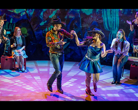 A man in a black cowboy hat, western shirt, jeans and cowboy boots dances with a woman in a cowboy hat, a skirt and cowboy boots as others watch on