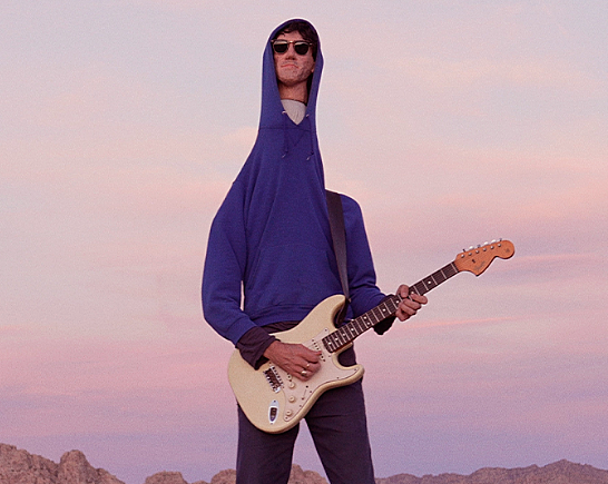 Delicate Steve wearing blue hoody playing pale yellow electric guitar with a desert sunset in the background