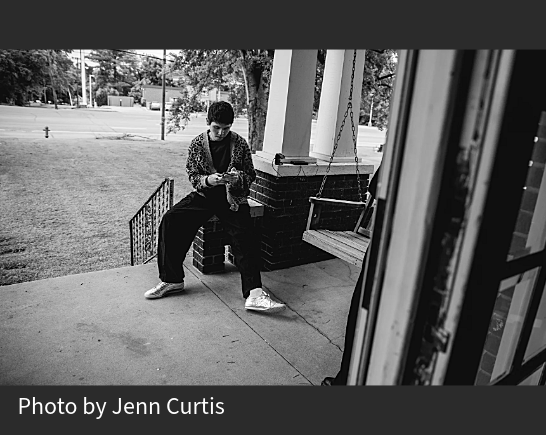 Musician Clinton Kane sits on a brick porch pier next to a wooden swing staring at a smart phone as the whole world moves on behind him.