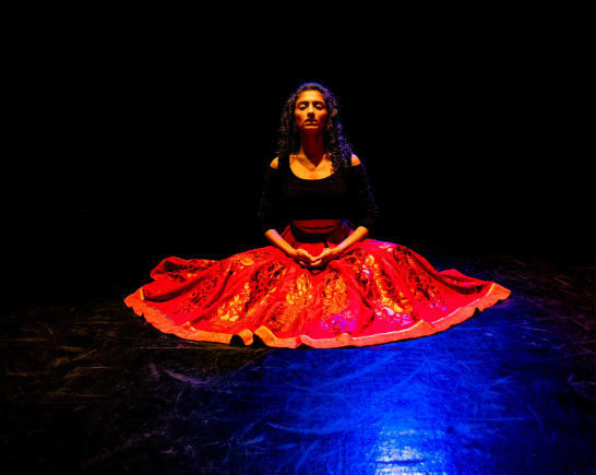 a dancer kneels on stage in a red skirt spread out about her, she wears a black top and has long wavy black hair. There is a blue pin spotlight shining on her, center stage