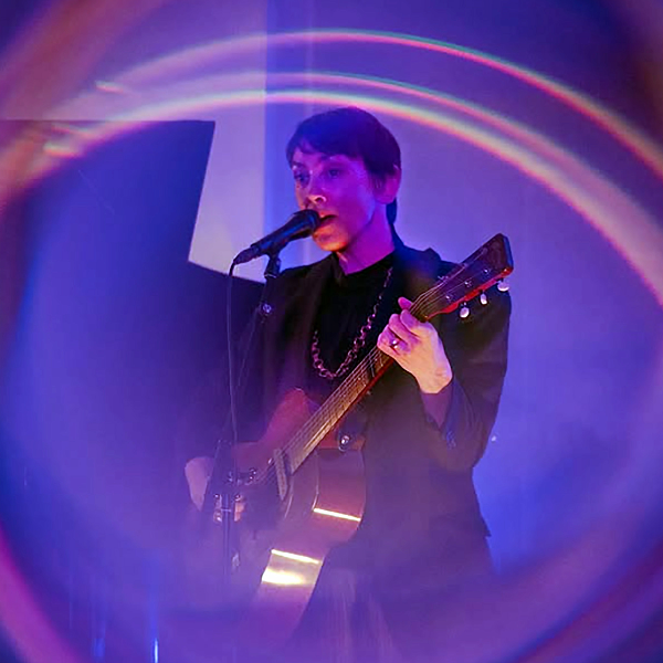 Woman with short brown hair plays an acoustic guitar and sings