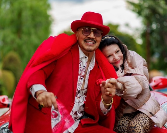 A red sports car parked with trees in the background while Actor Jesse Leigh who wears a bright red cape, suit and hat sits on the hood of the car with actress Irina Maleeva In a pink ski jacket and leopard print pants.