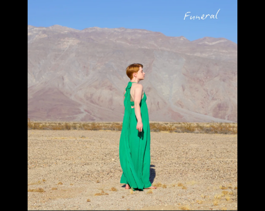 A fair skinned woman with auburn hair, wears a green sleeveless maxi dress as she stands in the desert with mountains in the background