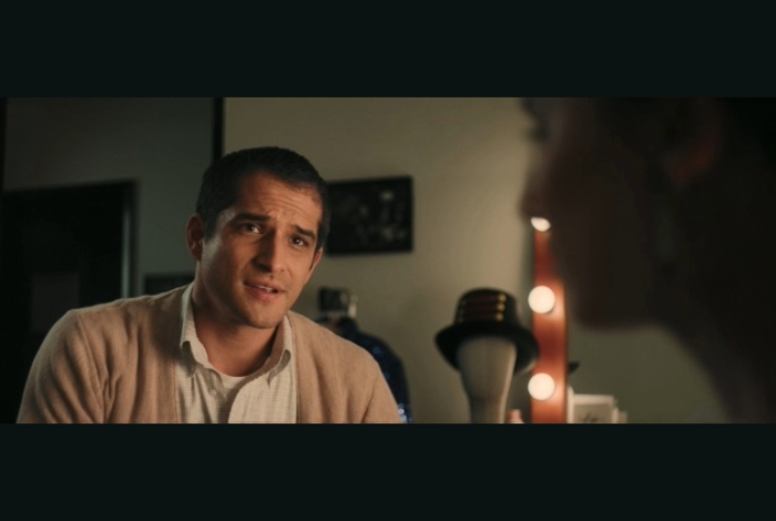 A still from the web series NADINE- a young man with short dark hair wears a beige sweater and white collared shirt. A dressing room mirror with lights is in the background as he faces the silhouette of a woman.