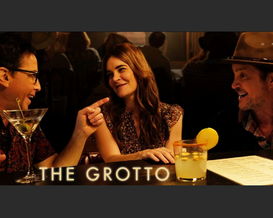 A young man with black framed glasses and short dark hair points and talks to a female who has long brown hair and wears a sleeveless shirt and man in a fedora type hat and short beard. The three of them sit at a bar with drinks in front of them.