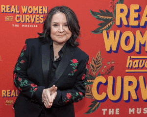 Woman with dark brown shoulder length hair wears a black silk suit with roses embroidered on it stands in front of a step-and-repeat for the Broadway Play Real Women Have Curves
