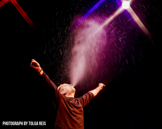 Actor Dan Ruth Stands center stage in a black box theatre, arms wide open spitting liquid into the air that is illuminated in the red tinted stage lighting