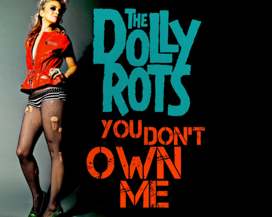 A blonde woman stands smiling next to words that say The DollyRots wearing a red sleeveless leather jacket with punk rock fashion zippers, she wears fishnet stockings with tears in them and green spike heel shoes.