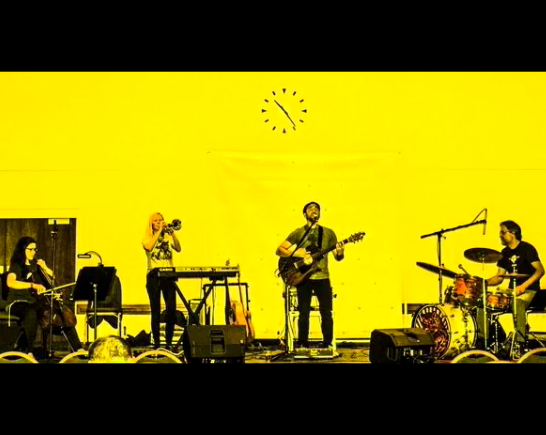 Four Musicians play on a stage with a yellow background with an abstract clock set at 10:24