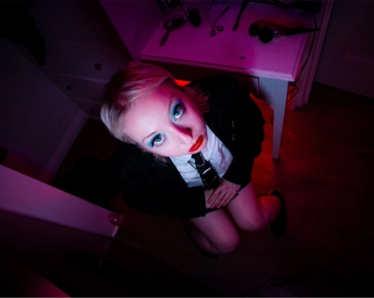 Music artist Kit major looks up wears a dark suit, white shirt, skinny black tie. She has blue eye shadow and red lipstick on