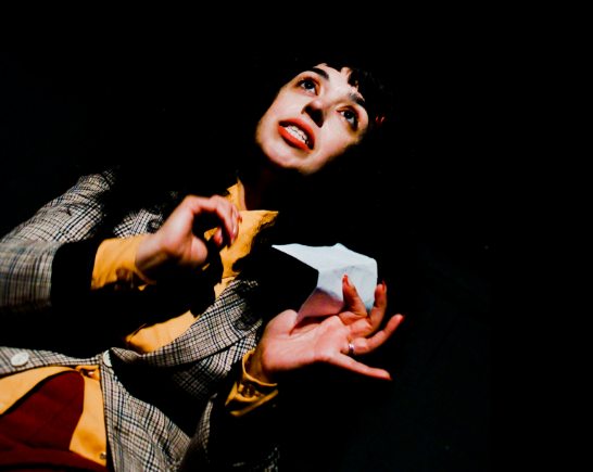 A woman on a dark stage stands in on spotlight, face turned to the light. She wears a plaid jacket, orange shirt and holds a piece of white paper in her hands... She has dark hair and wears red lipstick.