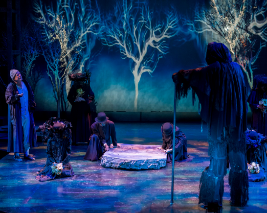 A darkly lit stage in purple and blue lighting. Bare trees upstage in a graveyard with 2 black clad spirits loom over two humans at graveside