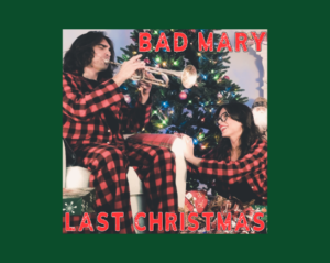 A man and a woman wearing matching plaid flannel pajamas sit in front of a christmas tree surrounded by presents