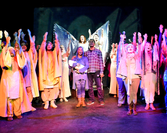 Onstage choir, with immigrant Mary, Joseph, baby Jesus and a Silver angel with wings spread