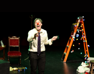 A man wearing a clown nose, a white button down shirt, tie and dark pants stands on a stage with a ladder draped in christmas lights. He laughs and points with his right arm
