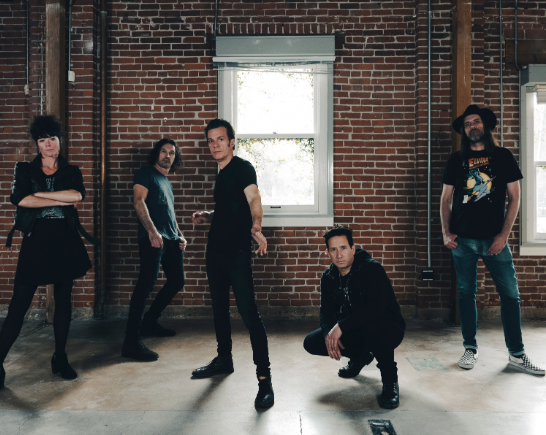Five people dressed in black stand on concrete floor of room with brick walls and large windows