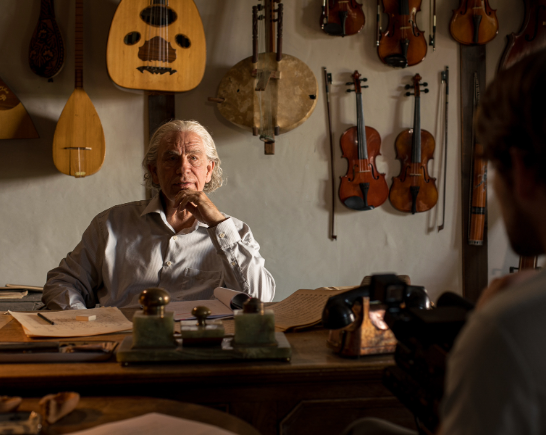 In the foreground a mans back faces a man with silver hair at a desk. His left hand rests on his chin. Multiple stringed instruments hang on the wall behind them.