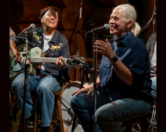 Two women sit on stools performing music. The band is The Graceland Exiles with Tina Schlieske on the left playing a guitar as her sister Laura Schlieske sings into a microphone