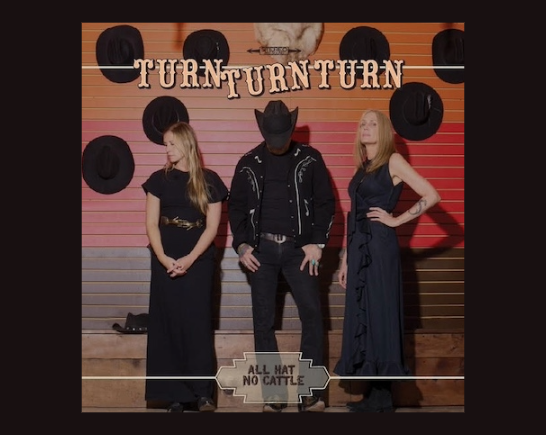 Black cowboy hats and a cow skull hang on an orange and tan wall. In front of the wall stands a man wearing a black cowboy hat, black western shirt with white stitching and black pants is flanked on each side by women with long blonde hair dressed in long black dresses