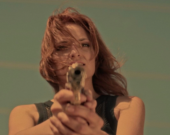 A woman with long windblown red hair holds a pistol with both hands pointing it straight ahead the desert sky behind her.