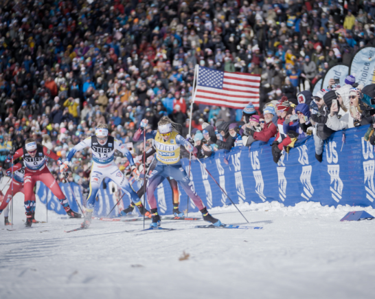 A crowd watches and cheers as Olympic Gold medalist, cross-country skier Jessie Diggins crosses the finish line carrying the American flag