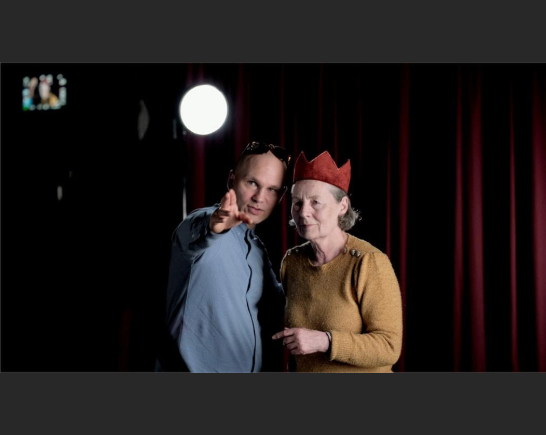 a frame from the documentary feature BRITA a man and woman stand center stage dressed in street clothes, she wears a red crown. His right arm is lifted straight ahead indicating forward to her