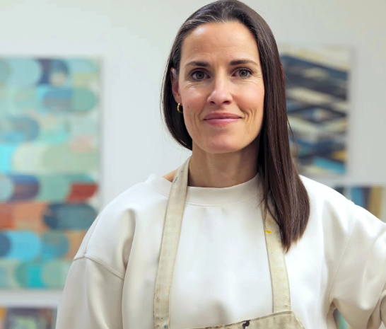 Artist Sunny Taylor a woman with long brown hair, wears a white crew neck shirt and a beige artists apron. Behind her are paintings from her exhibit CONNECTIONS