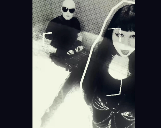 a man whose head is shaved sits in the far left corner of the image. He wears dark sunglasses and black clothing. A woman with dark black long hair wears black clothing and stands in the front right corner of the image