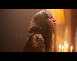 Side view of a woman with long dark tousled hair who is lit by candlelight, her face covered in blood