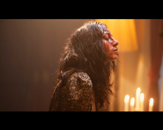Side view of a woman with long dark tousled hair who is lit by candlelight, her face covered in blood