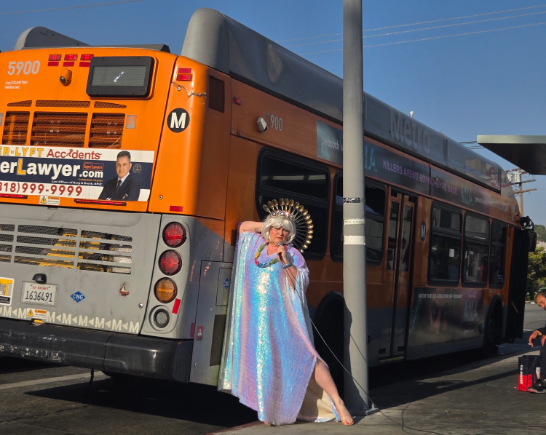 A woman in a blue rainbow colored gown wears a crown of spoons as she strikes a pose next to an orange Metro bus