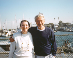 An elder couple smile warmly, arm in arm, in a marina with boats and harbor in the background