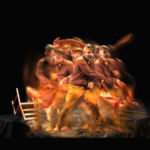 A man stands on a black stage surrounded by fire and wooden chairs turned askew