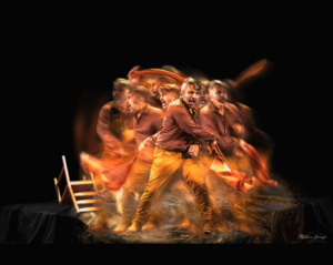 A man stands on a black stage surrounded by fire and wooden chairs turned askew