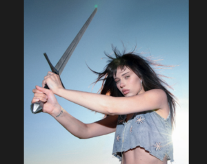 Music artist Thunder Bai, a pretty young woman with long dark hair, wears a light blue mid-drift top and wields a large silver sword. The pale blue sky with sunlight illuminate her from behind