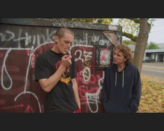 Two young men stand in front of a graffiti wall. The tallest one has a shaved head, wears a baggy tee shirt and lights a joint as the second young man is shorter with wavy blonde hair and wears a black sweatshirt. He has his hands in his pockets as he looks at the other young man as if questioning