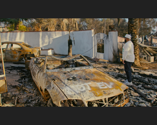 A man stands in the midst for the ruins of his burned home, the result of the Altadena Fire in Los Angeles. A burned out car with the words "Not EV" spray painted on the hood