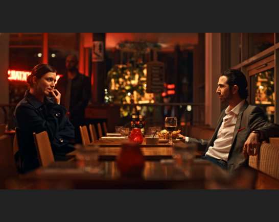 A restaurant table scene with a man in a suit leaning back with dominance as the woman across from him stares intensely at him as she bites her nails