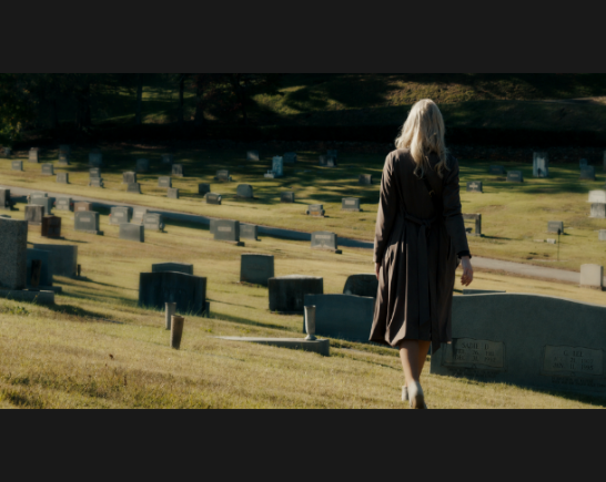 The back of a woman with long blonde hair, wearing a trench coat and heels walks through a cemetery with brown grass.