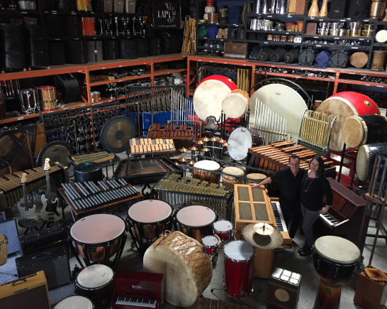 A man and woman stand inside a storage room full of percussion instruments