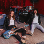 Rock duo Lips Speak Louder sit on the floor in front of a drum kit, one holds a blue guitar with a white pickup