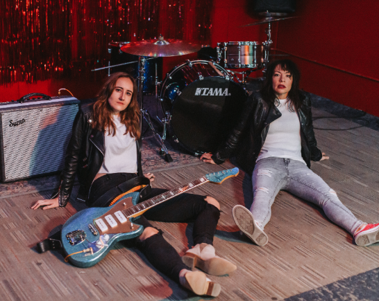 Rock duo Lips Speak Louder sit on the floor in front of a drum kit, one holds a blue guitar with a white pickup