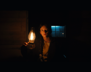 In a dark room a young woman with dark hair pulled back holds a kerosine lamp up so the glow of it covers the right side of her face