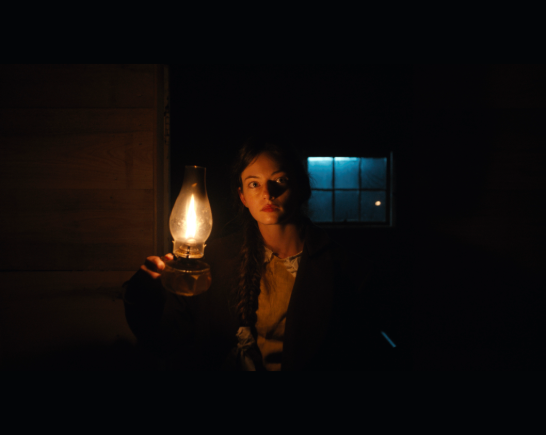 In a dark room a young woman with dark hair pulled back holds a kerosine lamp up so the glow of it covers the right side of her face