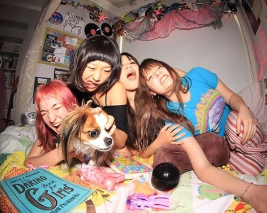 Four teenage girls members of the LA punk band Kim Theory are huddled together laughing with a cute dog and band fliers surrounding them