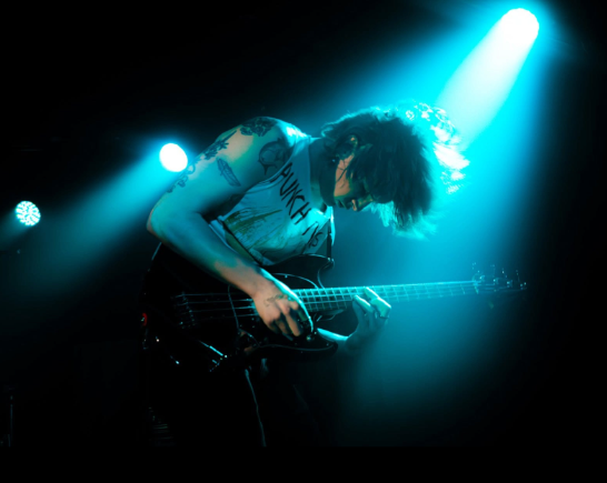 Guitar player from the band Trash Kickers leans over as he plays a black guitar in a blue/green spotlight on a dark stage