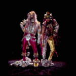 Performing artists Kaye Freeman and Amy Kaps stand on a black stage wearing handmade costumes, recycled materials, and theatrical interventions, Hibiscus TV